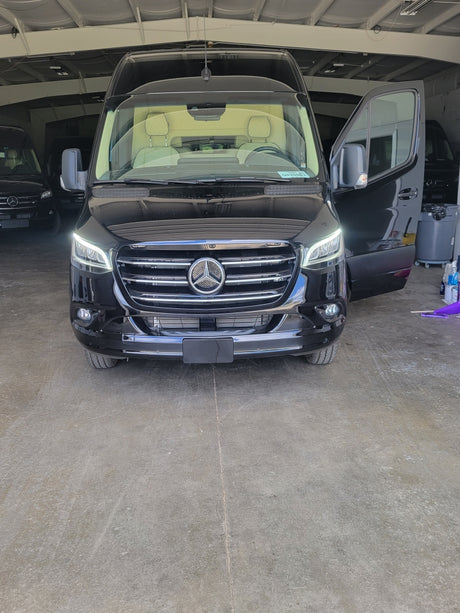Sleek black Mercedes-Benz Sprinter van for RV rental, headlights on, driver's door open, parked indoors.