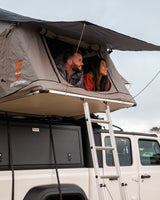 Happy couple smiles from their grey rooftop tent, mounted on a white **rv rental** vehicle, enjoying the outdoor adventure.