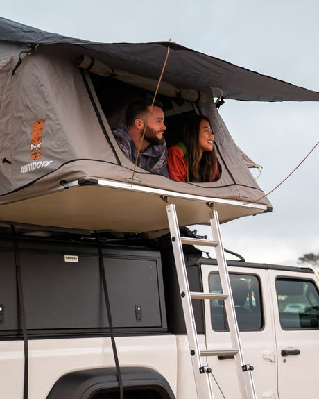 Happy couple smiles from their grey rooftop tent, mounted on a white **rv rental** vehicle, enjoying the outdoor adventure.
