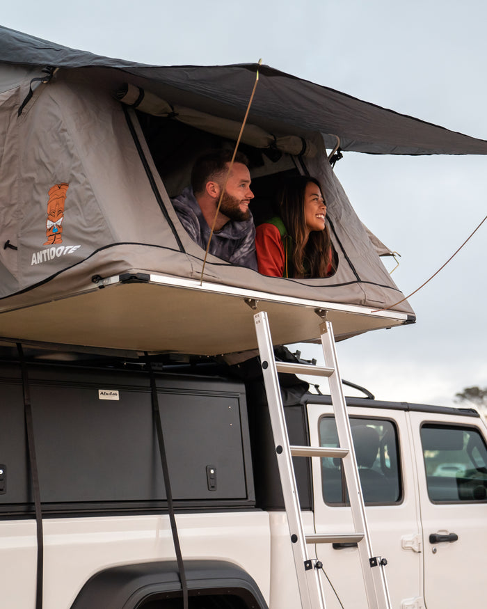 Happy couple smiles from their grey rooftop tent, mounted on a white **rv rental** vehicle, enjoying the outdoor adventure.
