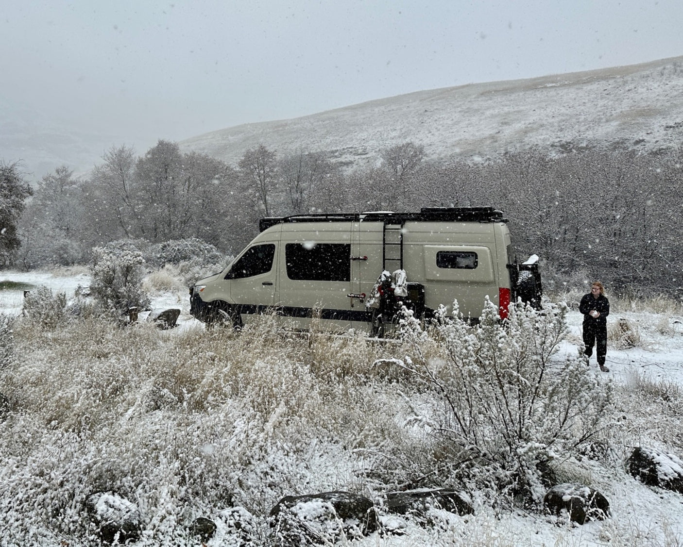 Beige campervan RV rental parked in a snowy winter landscape with a person standing nearby as snow falls lightly.