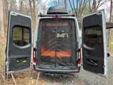 Rear of an RV rental van with doors open, revealing a bug screen and orange cushions inside, plus a Thule roof box.