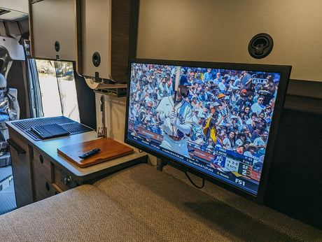 Inside an RV rental, a TV shows a baseball game next to a compact kitchen counter with a sink. Cozy sleeping area in foreground.