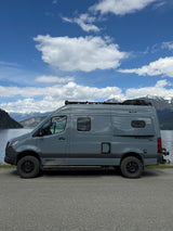 This grey Winnebago Revel 4x4 RV rental is parked by a stunning lake with snow-capped mountains and a blue sky.