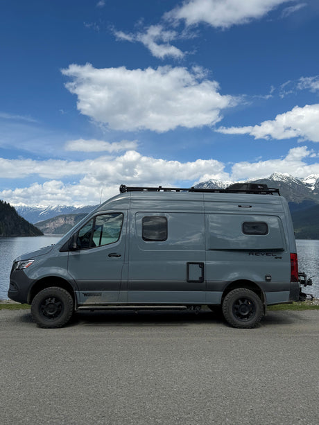 This grey Winnebago Revel 4x4 RV rental is parked by a stunning lake with snow-capped mountains and a blue sky.