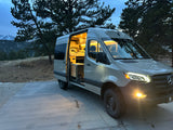 A modern **RV rental** camper van with an illuminated interior is parked at twilight, surrounded by trees and mountains. Perfect for adventure!