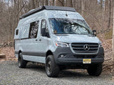 Light grey, lifted Mercedes-Benz Sprinter custom campervan with off-road tires, parked in woods. Ideal RV rental.