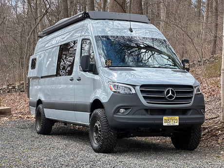 Light grey, lifted Mercedes-Benz Sprinter custom campervan with off-road tires, parked in woods. Ideal RV rental.