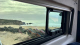 View from an RV rental window of a vast sandy beach, ocean waves, and rocky cliffs under a cloudy sky. People are on the beach.
