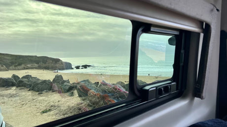View from an RV rental window of a vast sandy beach, ocean waves, and rocky cliffs under a cloudy sky. People are on the beach.