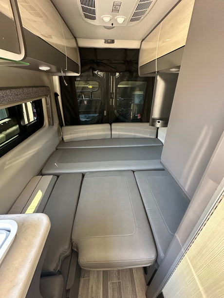 Interior view of a modern RV rental, featuring convertible light gray seating, overhead cabinets, and a side counter.