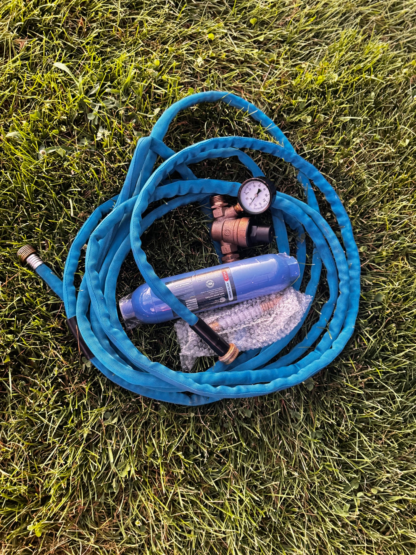 RV rental essentials: A blue RV water hose, inline water filter, and pressure regulator on grass, ready for safe camping.