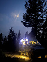 A brightly lit RV rental camper van parked at night under a moonlit sky in a dense forest. Cozy interior and outdoor setup.