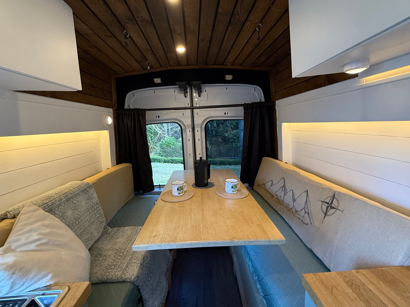 Cozy RV rental interior with a wooden dining table, comfortable bench seating, and scenic views out the back doors. Perfect for van life.