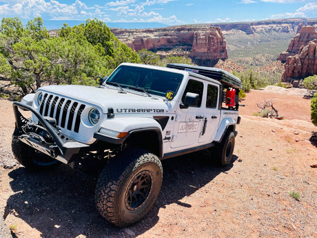A white Jeep Gladiator custom overland RV rental with a rooftop tent, set against a stunning desert canyon landscape.