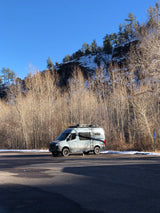 A Storyteller Overland camper van, like an RV rental, parked amidst bare trees and a snow-dusted hill.