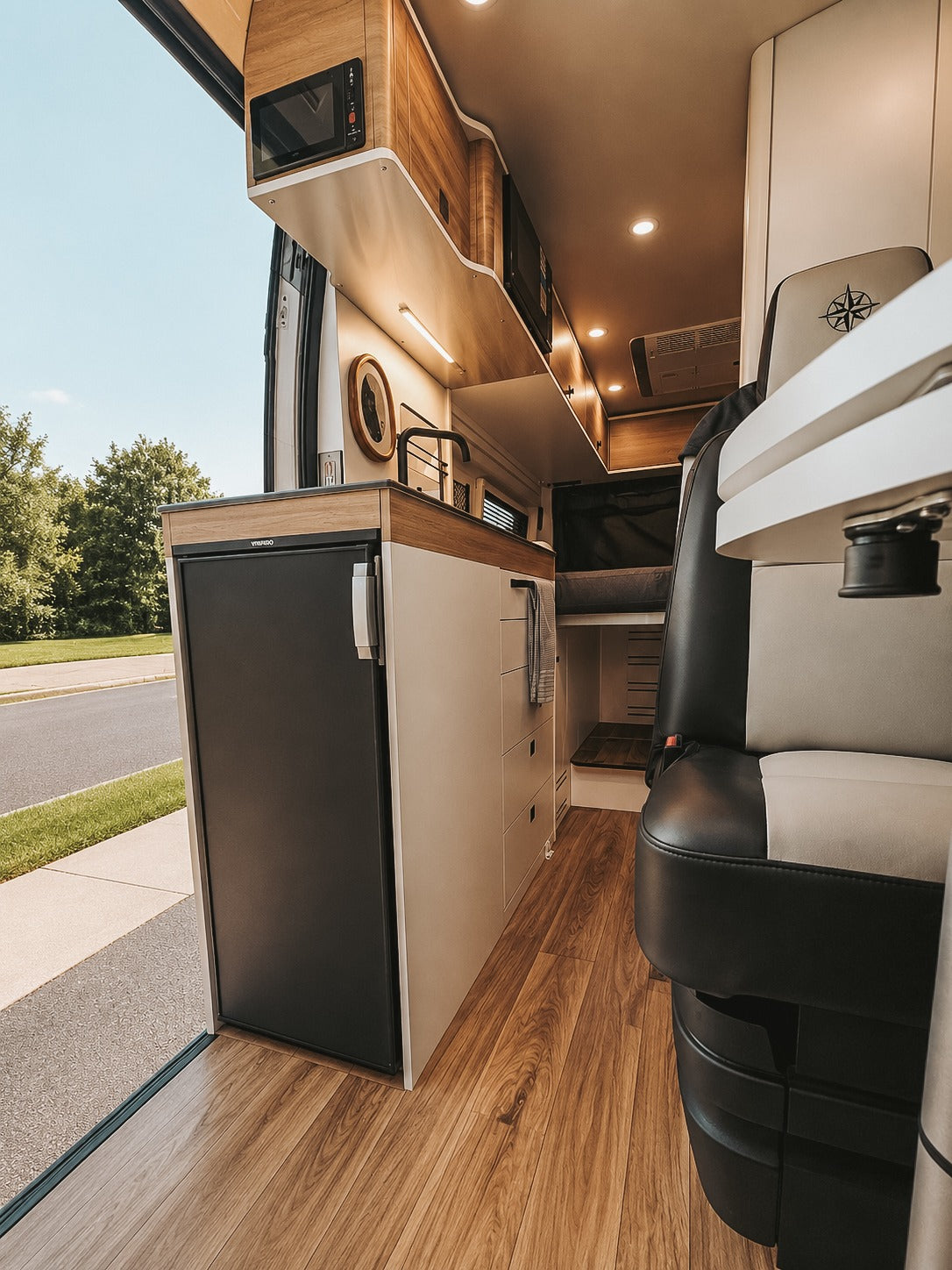 Modern rv rental interior with a sleek kitchenette (fridge, microwave, sink), wood floor, and open side door view. Ready for adventure.