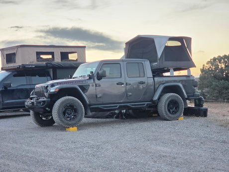 Grey Jeep Gladiator with a deployed roof top tent, like an RV rental, on a gravel campsite at sunset. Ready for overlanding adventures.