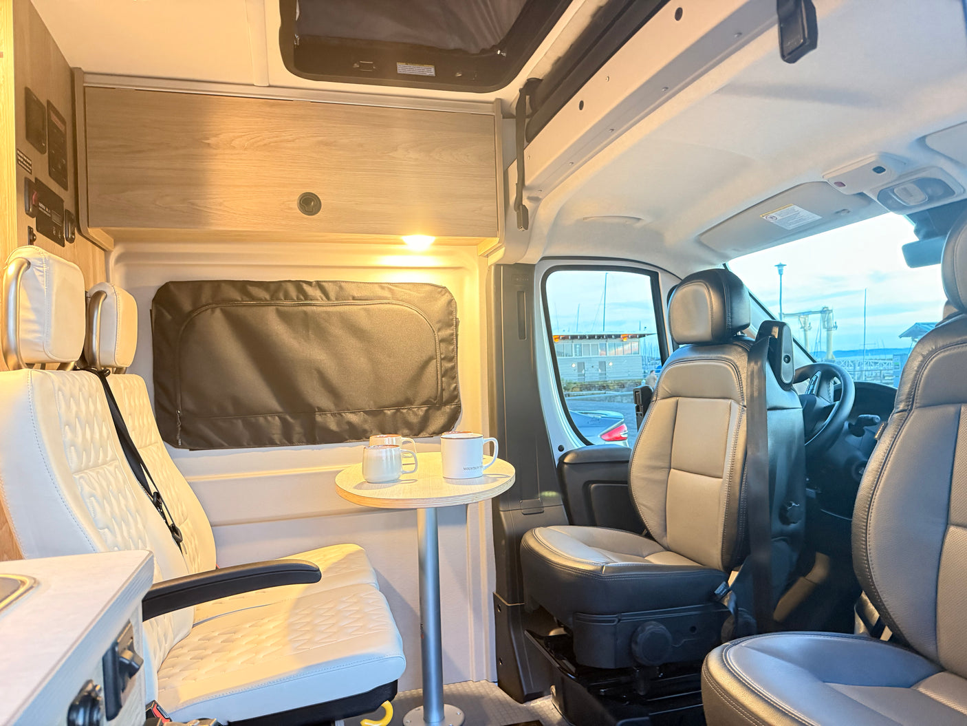 Bright RV rental interior showing white quilted seating, a small dining table with mugs, and light wood cabinets, ideal for travel.