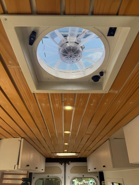 RV rental interior: Wooden plank ceiling, clear roof vent fan, recessed lighting, and practical white overhead storage cabinets.