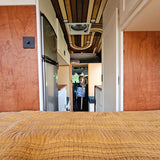 View inside an RV rental: a cozy bed in front, with a dog peering from the driver's area beyond custom wooden details.