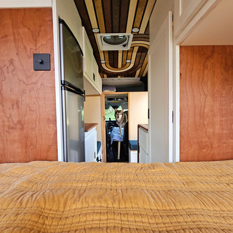 View inside an RV rental: a cozy bed in front, with a dog peering from the driver's area beyond custom wooden details.