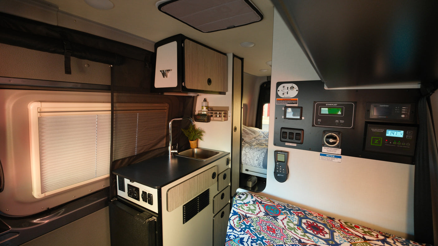 Inside this rv rental, a well-equipped camper van features a kitchen with sink, power control panel, and a patterned bed cushion.