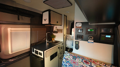 Inside this rv rental, a well-equipped camper van features a kitchen with sink, power control panel, and a patterned bed cushion.