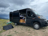 Dark grey Ram ProMaster campervan parked with doors open, showcasing an RV rental for van life. Solar panel, surfboard, & dog complete the scene.