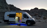 RV rental camper van with open illuminated kitchen, blue underglow, & bike rack, parked in a scenic desert landscape.