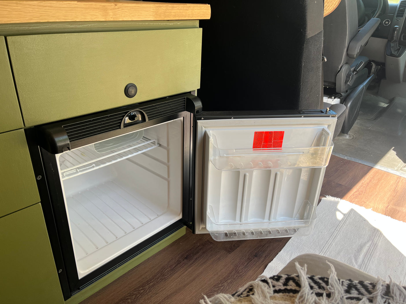 Open mini-fridge in an RV rental's olive-green camper van interior, showing clean shelves and door storage.