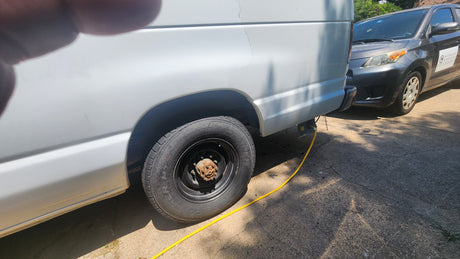 RV rental white van: Maintenance in progress. Rear wheel removed, exposing rusty hub. Yellow cable connected.
