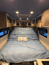Cozy RV rental camper van interior. A bed with blue bedding, striped pillow, overhead storage, lights, and side windows.