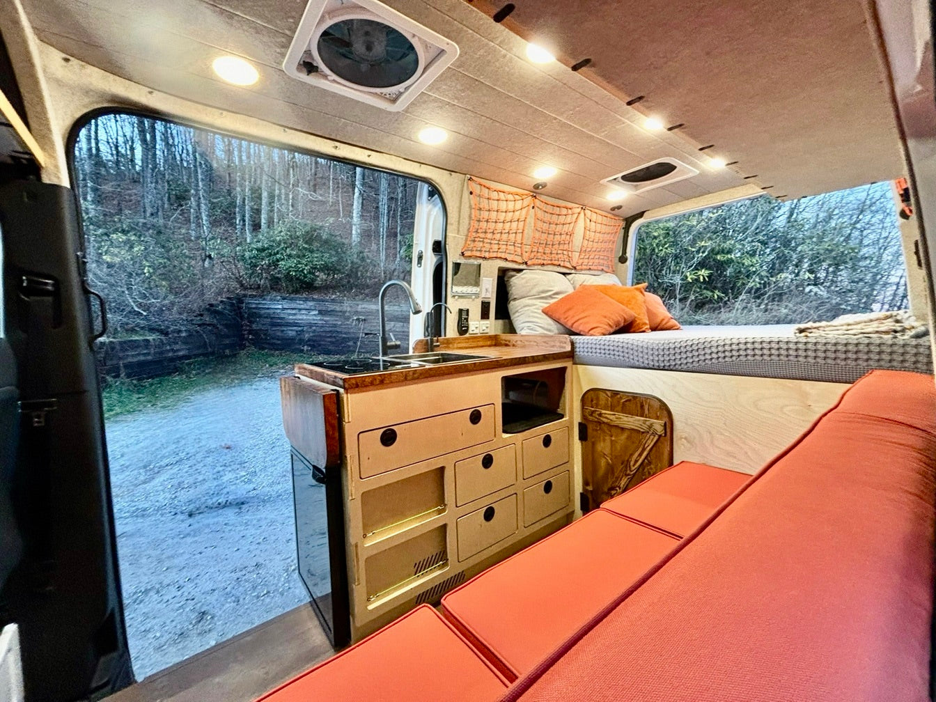 Inside a modern rv rental, showing a compact kitchen, cozy bed, convertible seating, and a forest view, perfect for mobile adventures.
