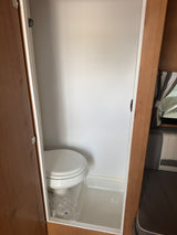 RV rental: Compact RV bathroom with a white toilet and integrated shower pan within a narrow, light-colored stall.