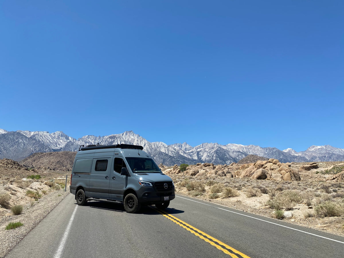 A gray Revel RV rental travels a scenic road with majestic snow-capped mountains in the background.