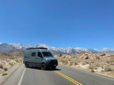 A gray Revel RV rental travels a scenic road with majestic snow-capped mountains in the background.