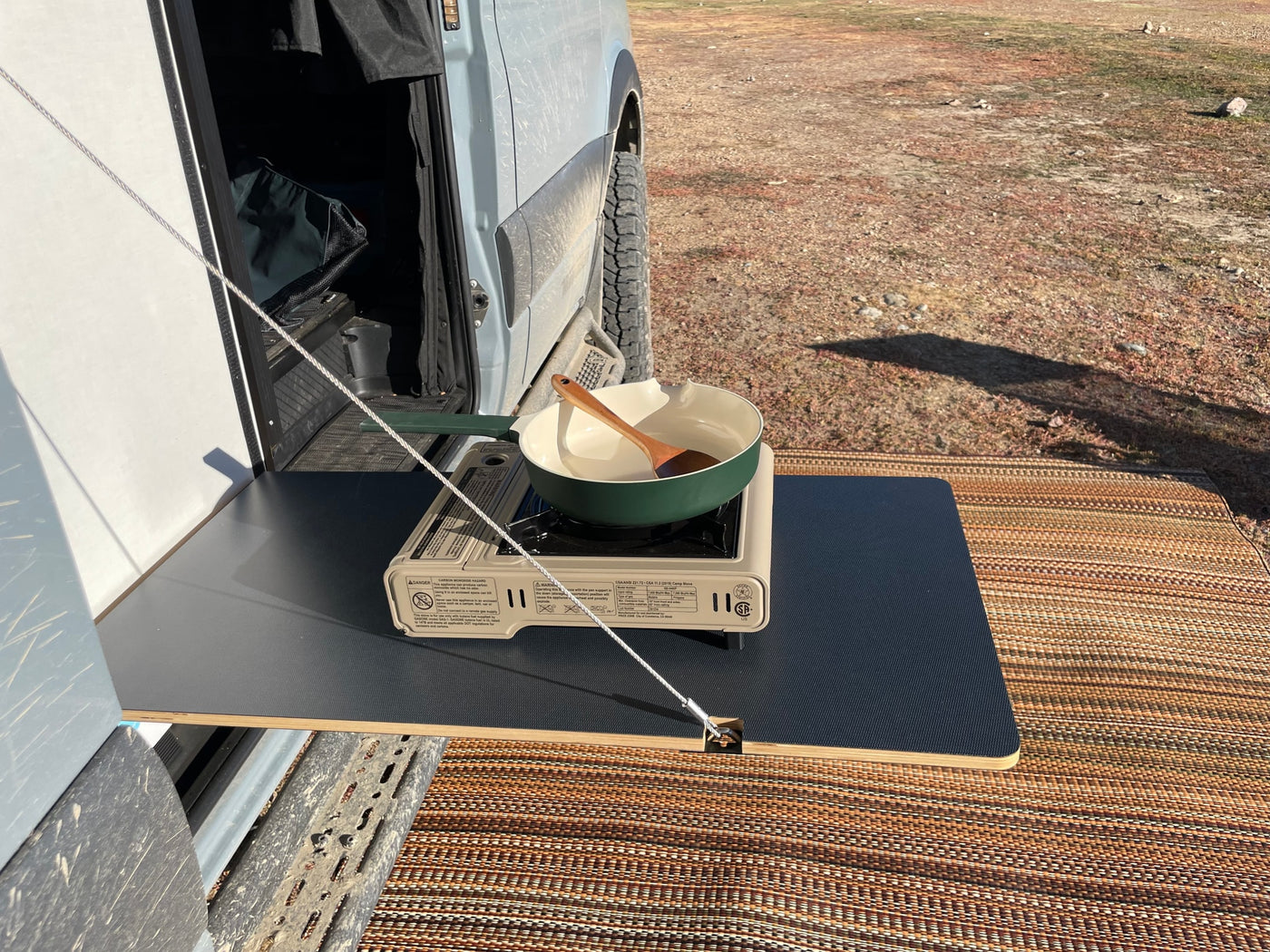 RV rental cooking setup: a portable stove, pot, and spoon on a pull-out table next to a camper van, perfect for outdoor meals. — photo 3