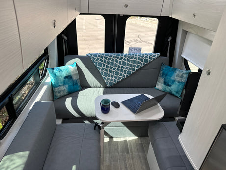 Cozy RV rental interior with grey seating, a white table with a laptop, and overhead cabinets, ideal for a mobile workspace.
