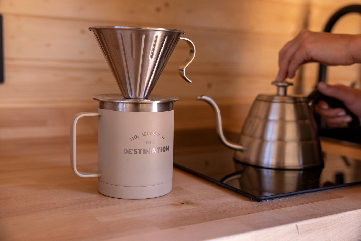 Hands pour hot water for pour-over coffee in an RV rental kitchen. A personalized mug and kettle are on a wooden counter.