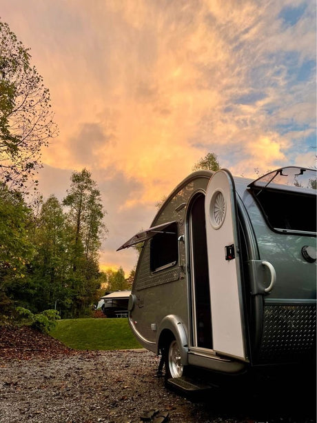 This image features a stylish gray RV rental teardrop trailer parked at a scenic campsite under a vibrant sunset sky.