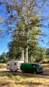 Green vintage van with silver camper, an ideal RV rental. Parked in a sunny field among tall trees.