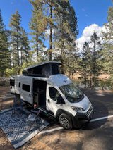 This RV rental camper van is perfectly set up for lakeside camping amidst tall pines, with chairs & an outdoor rug.