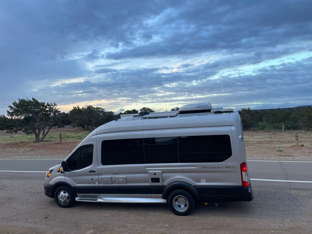 A silver Pleasure-Way campervan rv rental parked on a dirt shoulder by a road under cloudy skies, surrounded by trees.
