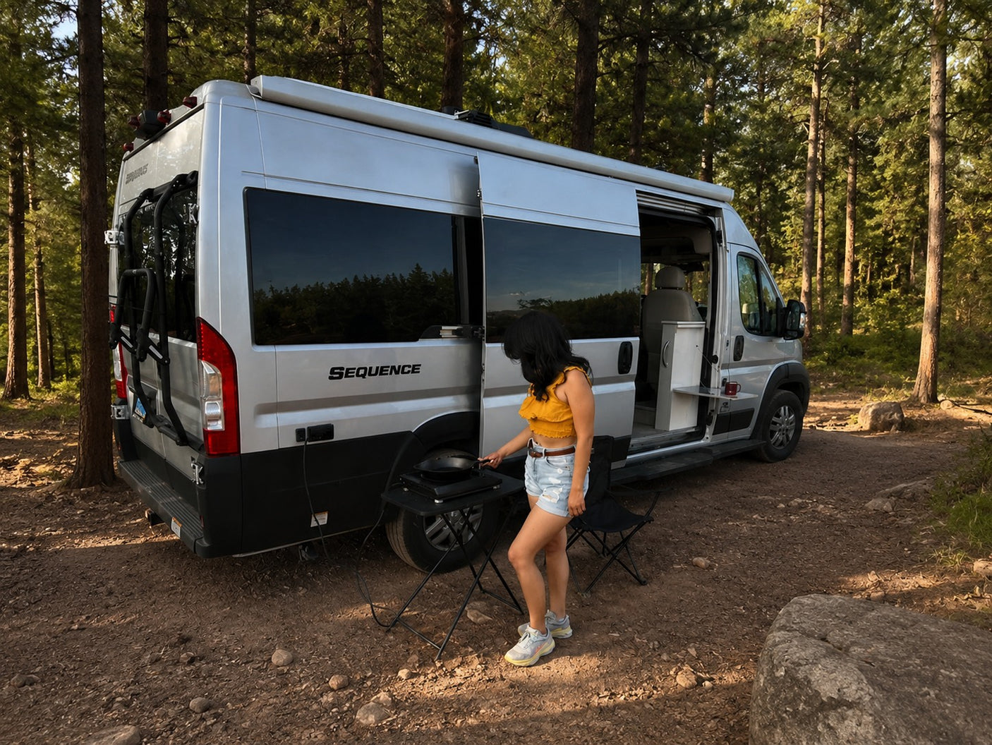 Enjoying an RV rental experience, a woman cooks outdoors next to a silver camper van in a serene forest setting.