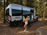 Enjoying an RV rental experience, a woman cooks outdoors next to a silver camper van in a serene forest setting.