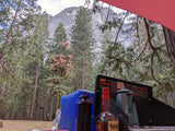 RV rental view: Campsite kitchen with a stove, coffee pot, and bottles. Tall pines frame a majestic mountain beyond.