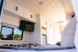 This bright RV rental interior features a cozy bed, wall-mounted TV, white cabinets, and a scenic window view.