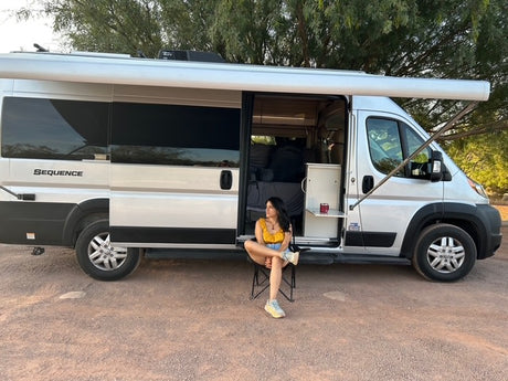Woman relaxes by her silver Sequence RV rental, awning extended, door open on a dirt road, offering a comfortable outdoor stop.