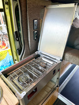 RV rental kitchen: Compact 2-burner stove and counter inside a campervan, with light switches and open door.